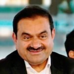 A smiling middle-aged man in a dark suit, with other people visible in the background, one of whom has a red tilak on their forehead