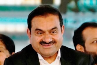 A smiling middle-aged man in a dark suit, with other people visible in the background, one of whom has a red tilak on their forehead