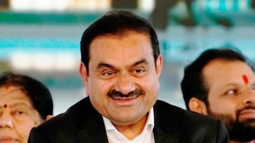 A smiling middle-aged man in a dark suit, with other people visible in the background, one of whom has a red tilak on their forehead