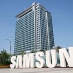 Large white Samsung logo letters installed outdoors in front of a tall modern corporate office building under a clear blue sky, with a worker walking nearby