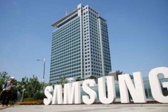 Large white Samsung logo letters installed outdoors in front of a tall modern corporate office building under a clear blue sky, with a worker walking nearby