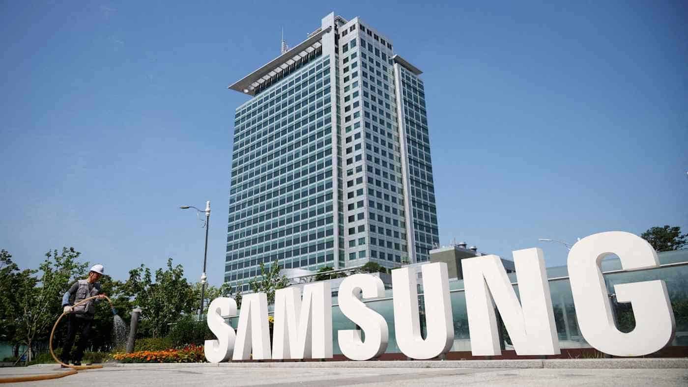 Large white Samsung logo letters installed outdoors in front of a tall modern corporate office building under a clear blue sky, with a worker walking nearby